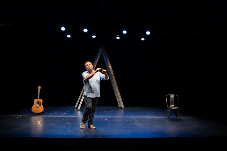 Au centre d’une scène, un homme habillé d’une chemise bleue et d’un jean gris, semble être en train de parler, il a les bras à demi levés, les index légèrement levés. Derrière lui, au fond de la scène, se trouve une échelle en fer. 